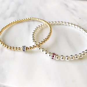 May include: Two beaded bracelets, one gold and one silver, with a small gemstone accent on each. The gold bracelet has a blue gemstone, and the silver bracelet has a red gemstone.