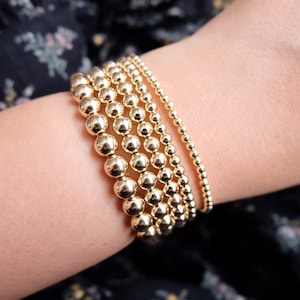 Op de afbeelding: Een verzameling gouden kralenarmbanden in verschillende maten. De armbanden worden om een pols gedragen en tonen de verschillende kraalmaten en de algehele glans van het goud. De armbanden zijn een eenvoudig maar elegant accessoire.