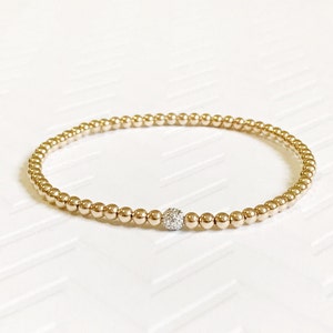 Pode incluir: Um bracelete de contas douradas com uma única conta maior no centro que é cravejada com pequenos diamantes.
