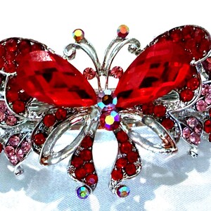 May include: A silver-toned metal hair clip shaped like a butterfly, featuring large faceted red crystals on the wings, smaller pink and red rhinestones, and iridescent accents. The design includes intricate metal filigree detailing throughout.