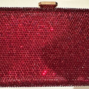Peut inclure: Une pochette rectangulaire rouge recouverte de petits ornements scintillants. La pochette a un fermoir doré et une surface solide et texturée. Le design général est glamour et accrocheur.