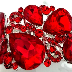 May include: A wide cuff bracelet featuring large, faceted, teardrop-shaped red crystals and smaller round red rhinestones set in a silver-toned metal frame. The design includes intricate, swirling metal patterns that connect the various crystal sizes.
