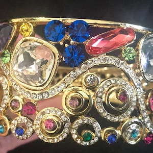 May include: Gold-toned cuff bracelet adorned with an array of colorful gemstones. The bracelet features large, clear, and colorful stones in various shapes, including blue, pink, and green, set against a backdrop of intricate gold filigree and smaller, sparkling crystals.