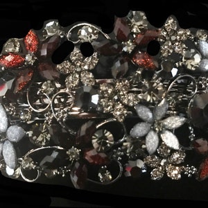 May include: An ornate hair comb featuring a floral design. The comb is embellished with sparkling rhinestones in shades of silver, gray, and burgundy. The flowers are arranged in a symmetrical pattern, creating a glamorous accessory.