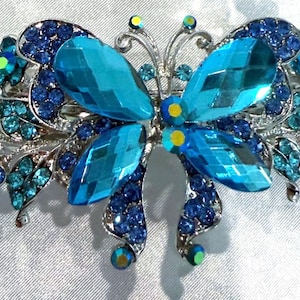 May include: A silver-toned hair clip shaped like a butterfly, featuring large faceted turquoise blue crystals for the wings and smaller dark blue and light blue rhinestones arranged in floral patterns across the metal frame.