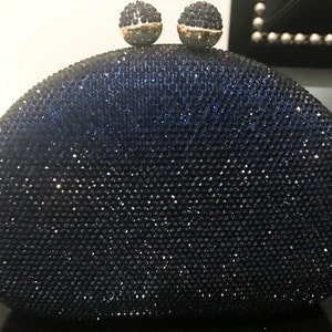 May include: A navy blue, oval-shaped clutch covered in small, sparkling embellishments. The clasp features two round, jewel-encrusted spheres with gold-colored bases. The bag is designed for evening wear and adds a touch of glamour.