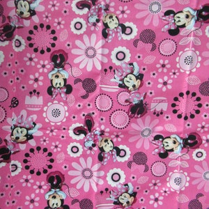 Puede incluir: Tela rosa con un patrón repetido de Minnie Mouse, flores y círculos. Minnie Mouse lleva un vestido rojo con lunares blancos y un lazo negro.
