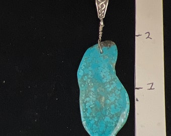 May include: A turquoise pendant with a silver bail. The pendant is an irregular shape, with a vibrant blue-green colour and a mottled pattern. The bail is a decorative silver piece with an ornate design. The pendant is suspended by a silver wire.