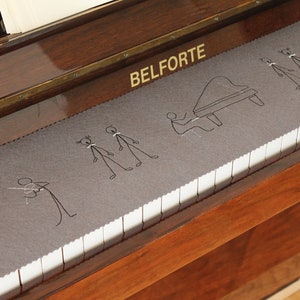 Peut inclure: Un piano avec une housse en feutre gris ornée de personnages brodés et d'un motif de piano. Les touches du piano sont blanches et noires. Le mot "BELFORTE" est visible sur le piano.