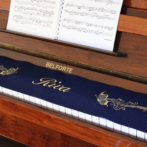 May include: A navy blue piano keyboard cover with gold embroidery of a butterfly and musical notes. The cover has the text "BELFORTE Riva" embroidered on it.