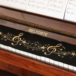 Puede incluir: Una funda negra para teclado de piano con notas musicales doradas, estrellas y la palabra "BELFORTE" bordada. El piano es de madera oscura y tiene teclas blancas. Partituras descansan sobre el atril.