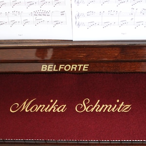 Può includere: Primo piano di un pianoforte in legno scuro con un top in feltro rosso. Il pianoforte è etichettato "BELFORTE" e ha un'iscrizione dorata che recita "Monika Schmitz".