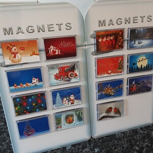 May include: Two white magnetic boards with rectangular Christmas-themed magnets. The magnets show festive images including snowmen, Santa Claus, Christmas trees, and holiday greetings. The word "MAGNETS" is printed at the top of each board.