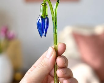 May include: A handcrafted glass flower sculpture featuring a vibrant blue, teardrop-shaped blossom with a bright green stem and leaves. The glass is smooth and translucent, capturing light through its delicate, elongated form and curved structure.
