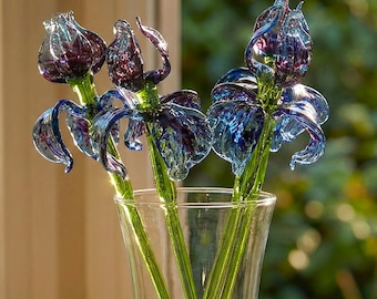 May include: A clear glass vase holds three glass iris flowers. The flowers have blue and purple petals and green stems. The vase sits on a wooden table with a book and a bowl.