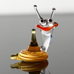 May include: A glass snail figurine with a clear body, orange and red mouth, and black eyes. The snail is sitting on a yellow glass shell.