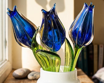 Peut inclure: Trois sculptures de tulipes en verre avec des pétales bleu profond et des tiges et feuilles vert translucide. Les fleurs sont disposées dans un vase cylindrique blanc à côtes. Les pétales en verre présentent de fines bulles.