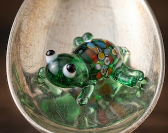 May include: A small green glass frog figurine with white and black eyes and a multicolored millefiori pattern on its back, resting inside the bowl of a tarnished silver-toned metal spoon with an ornate handle design at the base of the bowl.
