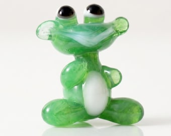 Puede incluir: Una pequeña figura de cristal verde translúcido con forma de rana. La criatura presenta un cuerpo liso y redondeado con una mancha ovalada blanca distintiva en el vientre, dos ojos negros prominentes y una boca ancha y sonriente con detalles blancos.