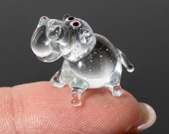 May include: A miniature clear glass figurine shaped like a hippopotamus. The small animal sculpture features a rounded body, four short legs, a short tail, and two dark red eyes. The glass is transparent with subtle internal air bubbles.
