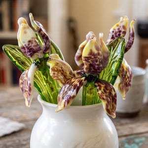May include: A collection of handcrafted glass flowers featuring intricate purple and cream mottled petals with vibrant green glass stems and leaves, arranged together in a smooth, white ceramic vase with a rounded base and narrow neck.
