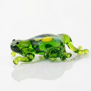 Puede incluir: Figurita de rana de cristal hecha a mano en tonos verdes, con detalles amarillos y azules en el dorso. La rana tiene detalles, incluyendo pequeños ojos negros y patas delicadas. Es un artículo decorativo.