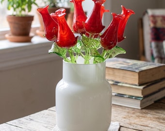 May include: A collection of vibrant red glass flowers with green glass stems and leaves arranged in a smooth, opaque white glass vase. The flowers feature elongated, tulip-like petals with a glossy finish, standing upright within the cylindrical vessel.