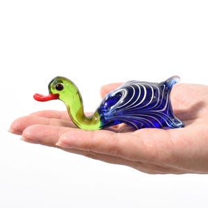 May include: A small glass duck figurine featuring a translucent light green neck and head with a bright red beak. The body is crafted from deep blue glass with intricate, swirling white marbled patterns throughout the rounded, tail-up shape.
