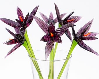 May include: A collection of five handcrafted glass flowers with deep purple, translucent petals and bright orange centers. Each flower is attached to a long, slender, translucent green glass stem, and the entire arrangement is displayed in a clear glass vase.