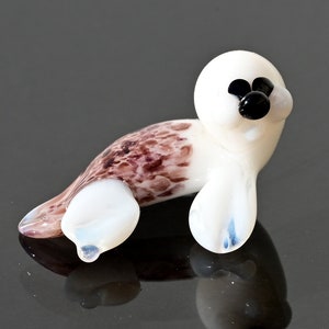 May include: A small, glass seal figurine. The seal has a white head and flippers, with a brown and white spotted body. The eyes and nose are black. The figurine is resting on a reflective surface.