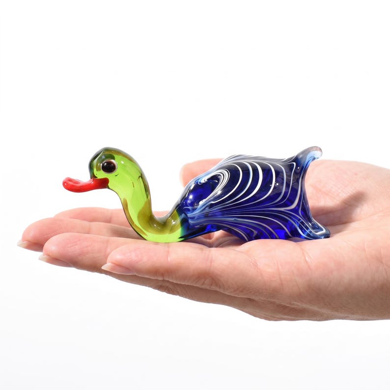 May include: A small glass duck figurine featuring a translucent light green neck and head with a bright red beak. The body is crafted from deep blue glass with intricate, swirling white marbled patterns throughout the rounded, tail-up shape.