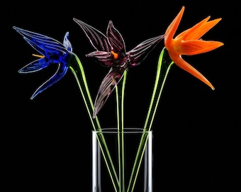 Puede incluir: Tres flores de cristal de colores en un jarrón transparente sobre un fondo negro. Las flores son azules, moradas y naranjas, con largos tallos verdes. El jarrón es cilíndrico y transparente.