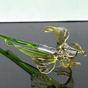 May include: A glass flower sculpture with a yellow flower and green stem. The flower is made of blown glass and has a delicate, flowing design.