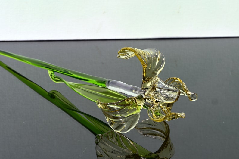 May include: A glass flower sculpture with a yellow flower and green stem. The flower is made of blown glass and has a delicate, flowing design.