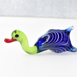 May include: A small glass figurine shaped like a duck. The head and neck are light green with a bright red beak and dark eyes. The body is a deep blue glass featuring a swirling white marbled pattern throughout the rounded, tail-tipped form.