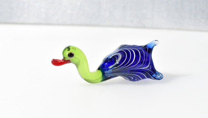 May include: A small glass figurine shaped like a duck. The head and neck are light green with a bright red beak and dark eyes. The body is a deep blue glass featuring a swirling white marbled pattern throughout the rounded, tail-tipped form.