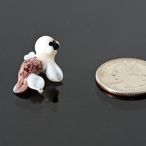 May include: A miniature glass seal figurine with a white head and flippers, a brown and purple body, and black eyes. A US quarter coin is next to the seal for size comparison.