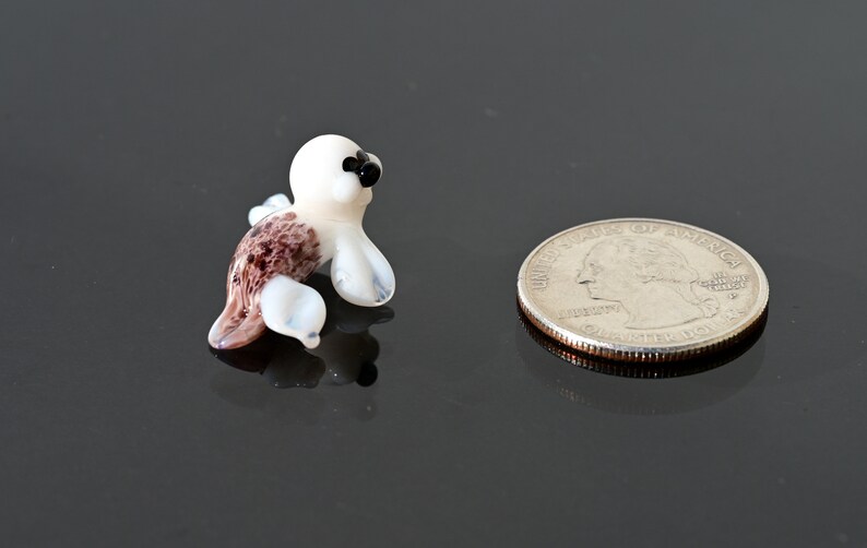 May include: A miniature glass seal figurine with a white head and flippers, a brown and purple body, and black eyes. A US quarter coin is next to the seal for size comparison.