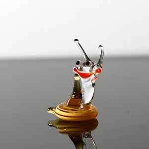 May include: A glass snail figurine with a smiling face, red and orange accents, and a golden shell. The snail is standing on a reflective surface.