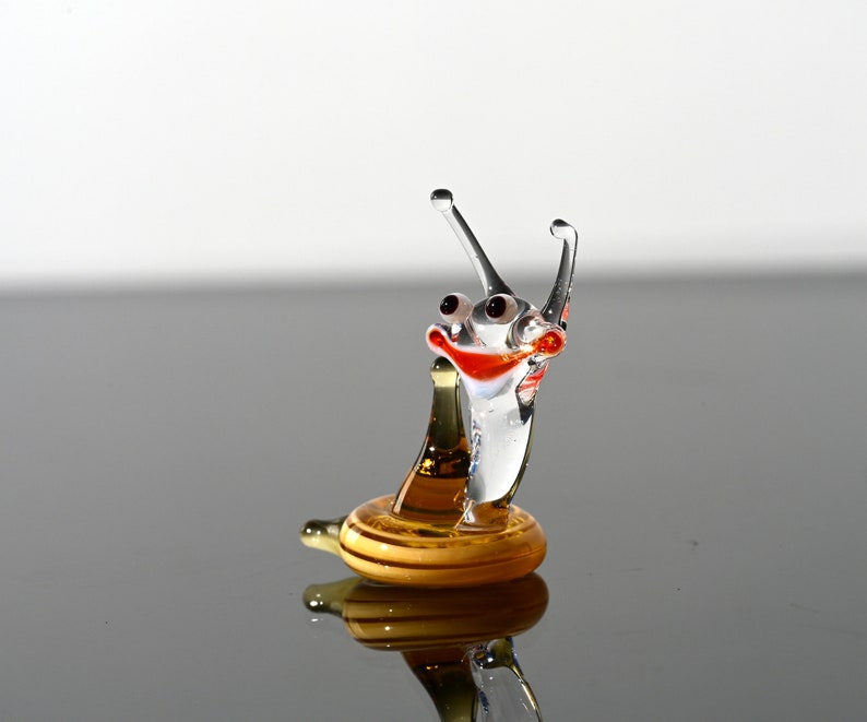 May include: A glass snail figurine with a smiling face, red and orange accents, and a golden shell. The snail is standing on a reflective surface.