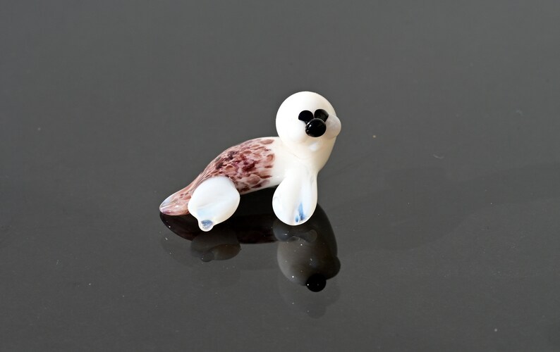 May include: A small, glass seal figurine. The seal has a white head and flippers, with a brown and white spotted body. The eyes and nose are black. The figurine is resting on a reflective surface.
