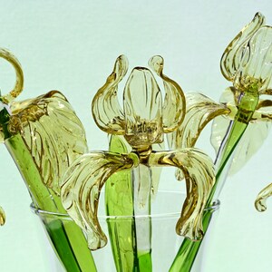 May include: Three glass iris flowers in a clear vase. The flowers are a light yellow color and have green stems.