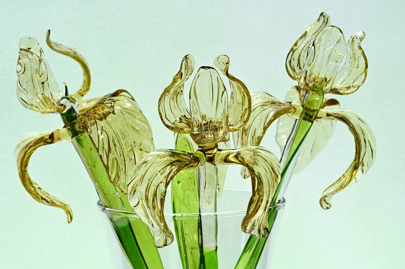 May include: Three glass iris flowers in a clear vase. The flowers are a light yellow color and have green stems.