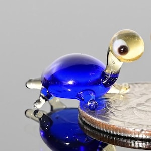 Peut inclure: Une figurine de tortue en verre miniature dotée d'une carapace bleu cobalt vif et d'une tête ambrée claire translucide. La tortue possède un œil brun foncé visible et des membres en verre transparent, posée sur le bord d'une pièce métallique.