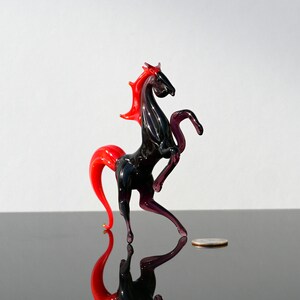 Peut inclure: Une figurine de cheval en verre noir avec une crini&egrave;re et une queue rouges. Le cheval est debout sur ses pattes arri&egrave;re, les pattes avant lev&eacute;es. La figurine mesure environ 10 cm de haut.