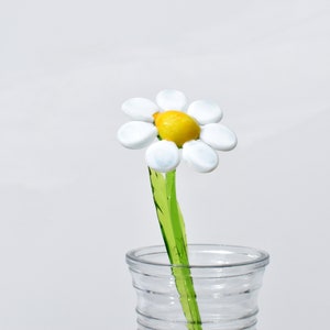 May include: A white glass daisy flower with a yellow center, on a green stem, in a clear glass vase.