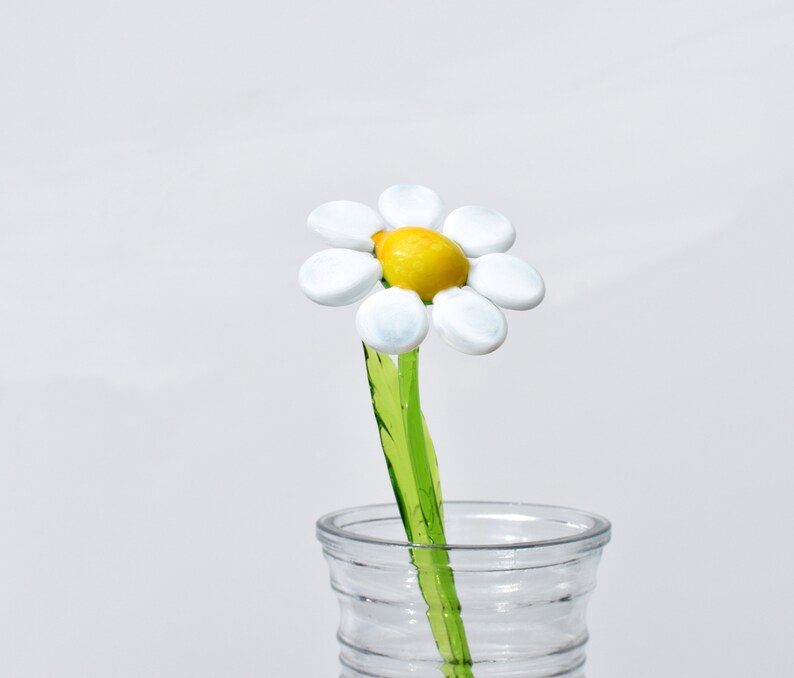 May include: A white glass daisy flower with a yellow center, on a green stem, in a clear glass vase.