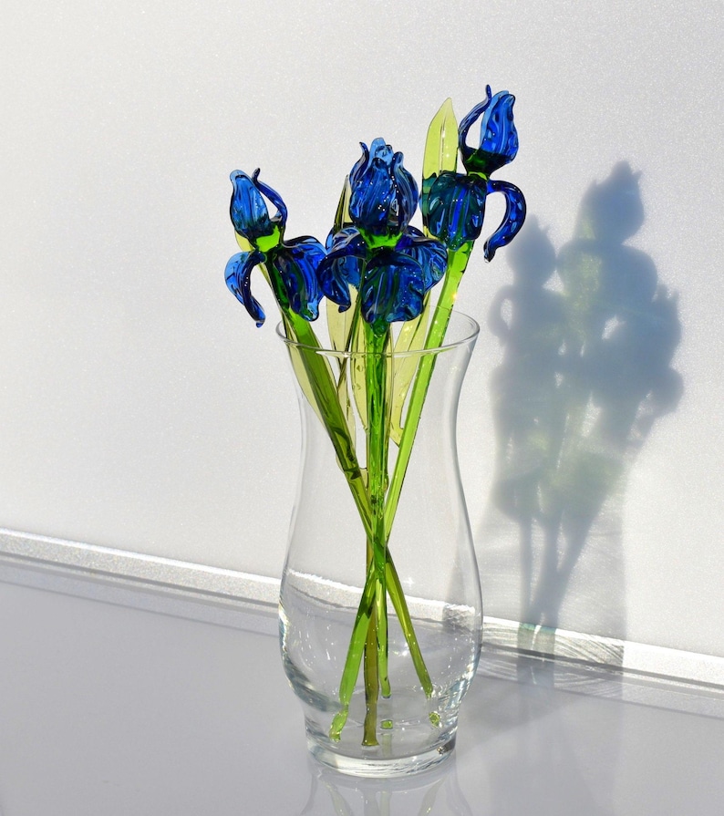 May include: A clear glass vase with a round base and a wide mouth. The vase contains five blue glass iris flowers with green stems. The flowers are arranged in a bouquet and are facing different directions.