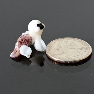 May include: A miniature glass seal figurine. The seal has a white head and flippers, with a brown and white spotted body. The seal is next to a U.S. quarter coin for size comparison.