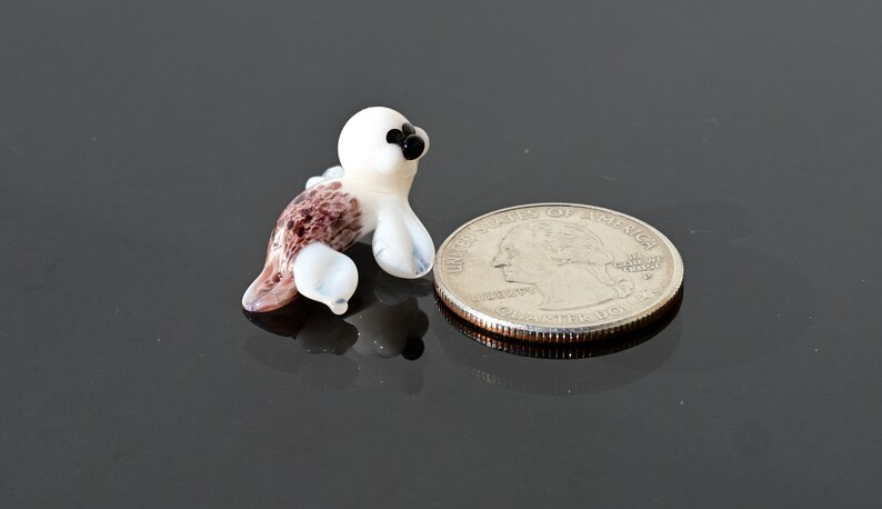 May include: A miniature glass seal figurine. The seal has a white head and flippers, with a brown and white spotted body. The seal is next to a U.S. quarter coin for size comparison.